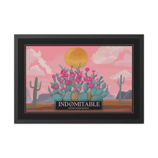 A framed art print featuring a stylized desert landscape with a large yellow sun, pink sky, blooming green cacti with pink flowers, and a black sign reading 'INDOMITABLE' at the bottom. The print is housed in a black frame and shown against a plain white background.