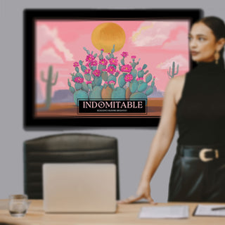 A framed art print featuring a stylized desert landscape with pink cacti, a yellow sun, and the text 'INDOMITABLE' is displayed on a wall in an office setting. A woman in a black sleeveless top and black skirt stands beside a desk with a laptop.