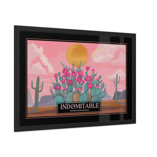 Framed art print featuring a stylized desert landscape with a blooming cactus, pink sky, clouds, and a large yellow sun. The print includes the text 'INDOMITABLE' resilience blooms brightest, on a black label at the bottom, shown in a black frame against a white background.